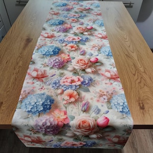 May include: Rectangular floral table runner with a watercolour design of roses, hydrangeas, and other flowers in pink, blue, and peach. The runner rests on a wooden table. A decorative accent for dining or home decor, measuring approximately 150 cm in length.