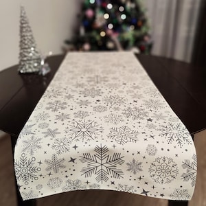 May include: A white table runner with a snowflake pattern in grey. The runner is on a dark brown table.