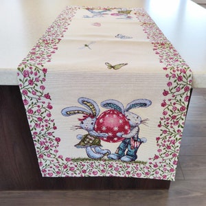 May include: A white table runner with a pink floral border. The center features two bunnies holding a large red and white Easter egg. The design is surrounded by a pink floral border.