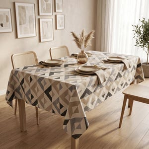 May include: A rectangular tablecloth featuring a geometric pattern of diamonds and triangles in shades of beige, gray, and white. The fabric covers a wooden dining table set with ceramic plates, gold-toned cutlery, and a glass vase with dried grass.