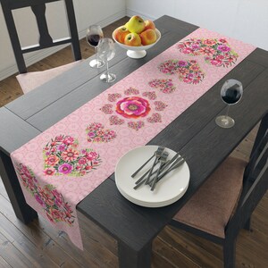 Puede incluir: Un camino de mesa rosa con un diseño floral con corazones y flores. El camino de mesa está sobre una mesa de madera oscura con platos blancos y copas de vino.