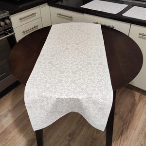 May include: A light grey table runner with a white floral pattern, draped over a dark brown round table. The runner's design features swirling, symmetrical motifs. The table is in a kitchen setting with white cabinets and a wooden floor.