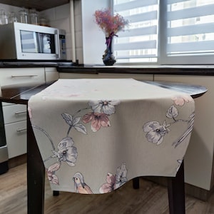 May include: A light green table runner with a floral pattern featuring pink and white flowers and a small bird. The runner is on a dark brown wooden table.