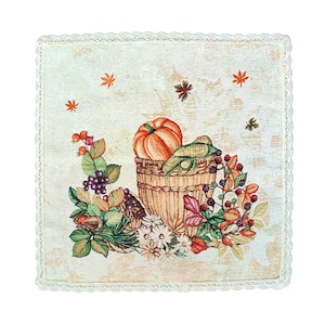 Puede incluir: Un mantel individual cuadrado de tela de color crema con un borde de encaje blanco. El centro presenta una escena de cosecha de otoño colorida con una calabaza, bayas, hojas y una cesta.