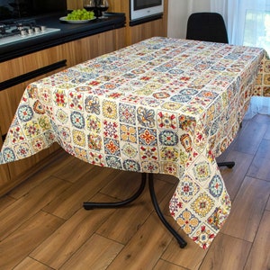 May include: A white tablecloth with a colorful geometric pattern featuring squares, circles, and floral designs. The tablecloth is draped over a black metal table with a wooden floor in the background.