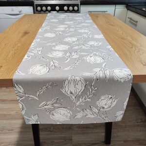 May include: A grey table runner featuring a floral design of white flowers and black outlines. The runner is laid on a wooden table with black legs. The floral pattern is repeated across the fabric.