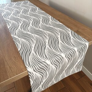 May include: A white table runner with a grey wavy pattern. The runner is on a wooden table.
