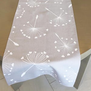 May include: A grey table runner with a white dandelion pattern. The dandelion design features heart-shaped seeds and delicate stems, creating a soft, nature-inspired aesthetic. The runner is made of a soft fabric.