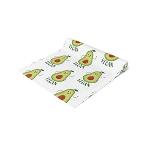 May include: White table runner with a repeating pattern of green avocados with smiling faces and the word "VEGAN" printed in green.