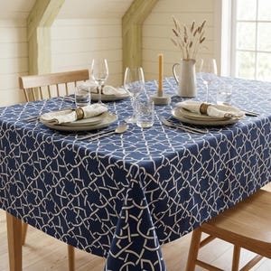 May include: A navy blue tablecloth with a white geometric interlocking pattern covering a dining table. The table is set with plates, glasses, silverware, cloth napkins with gold rings, a tall candle in a holder, and a vase with dried grass.