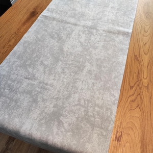 May include: A pale grey table runner with a textured, mottled design. The rectangular runner is placed on a wooden table. The fabric of the runner looks soft and has a slight sheen, adding a touch of elegance to the table setting.