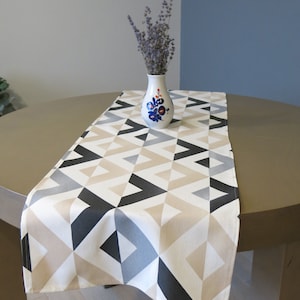 May include: A beige and black geometric table runner with a chevron pattern. The runner is on a brown table with a white vase of lavender flowers.