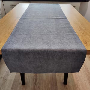 May include: A grey table runner made from a soft, textured fabric. The runner is placed on a wooden table with black legs. The table is in a well-lit room, and the runner adds a touch of elegance to the table setting.