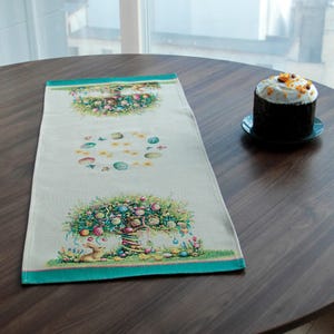 以下が含まれることがあります： 淡いベージュの背景とターコイズの縁取りが施された、長方形のイースターをテーマにしたテーブルランナー。ランナーは、カラフルなイースターエッグとリボンで飾られた木のデザインが特徴です。白いフロスティングを施した焼き菓子が、濃い色の皿の上に置かれています。