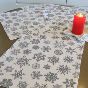 May include: Three white placemats with a snowflake pattern in grey. Two red candles are on the placemats, each on a white star-shaped plate.