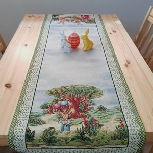 May include: A green and white table runner with a lace border features a woven design of bunnies, balloons, and a tree. The runner is decorated with three bunny figurines and a decorated Easter egg.