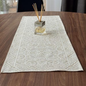 Puede incluir: Un camino de mesa rectangular beige con un patrón detallado y repetido está centrado en una mesa de madera oscura. Un difusor de vidrio con varillas de madera y una parte superior similar al hormigón se encuentra en el camino.