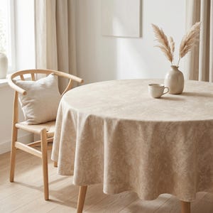 May include: A round beige tablecloth on a wooden table, with a matching chair and cushion. A vase with dried plants and a small cup are on the table. The room has neutral curtains and a minimalist design. The table is approximately 120 cm in diameter.