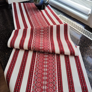 May include: White table runner with red and black embroidered floral patterns and vertical stripes. The runner is folded over, showcasing the detailed design. The runner is made of fabric.