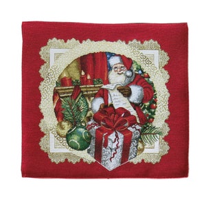 以下が含まれることがあります： 白いレースの縁取りが付いた赤いクリスマスのテーブルランナー。中央には、暖炉のそばでリストを読んでいるサンタクロースと、ギフトボックスとオーナメントが描かれています。