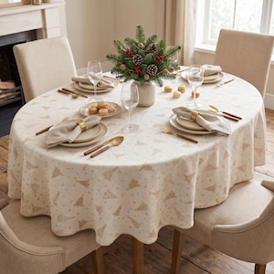 Op de afbeelding: Een ovale eettafel bedekt met een crèmekleurig tafelkleed met een goudkleurig kerstboompatroon. De tafel is gedekt met witte borden, gevouwen servetten met gouden ringen, wijnglazen, goudkleurig bestek en een tafelstuk in het midden.