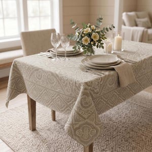 May include: A beige tablecloth with a paisley pattern covers a wooden dining table set for a meal. The table is set with white plates, silverware, wine glasses, and a floral centerpiece. A beige chair is behind the table.