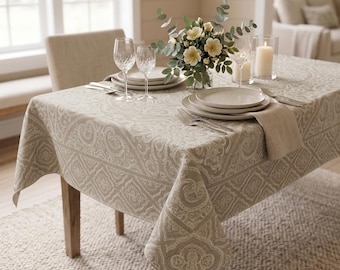 Puede incluir: Una mesa de comedor de madera cubierta con un mantel beige con estampado de cachemira. La mesa está puesta con platos blancos, cubiertos, copas de vino y un centro de mesa floral. Una silla beige está detrás de la mesa.