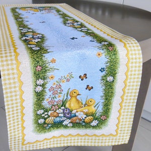 May include: A yellow and white table runner with a scalloped edge. The centre of the runner features a blue background with a green grass border and a scene of two yellow ducklings surrounded by flowers and Easter eggs.