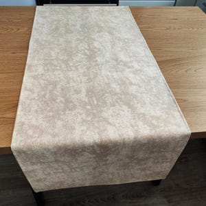 May include: A beige table runner with a textured, mottled design. The rectangular runner is laid on a wooden table. The neutral colour makes it suitable for a variety of interior design schemes.