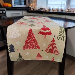 May include: A beige Christmas table runner with a repeating pattern of red, green, and white Christmas trees and snowflakes. The trees vary in design, with some detailed and others more simple. The runner is placed on a dark wooden table.