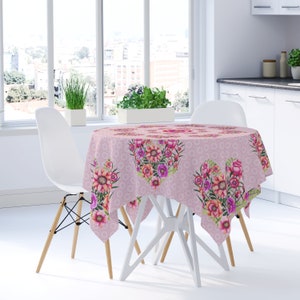 May include: Pink round tablecloth with a floral pattern featuring pink, orange, and purple flowers. The tablecloth is laid on a white table with two white chairs.