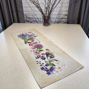 以下が含まれることがあります： ベージュのテーブルランナーで、紫、ピンク、白、黄色の花が様々な種類咲いています。花はランナーの中央に縦に並んでいます。
