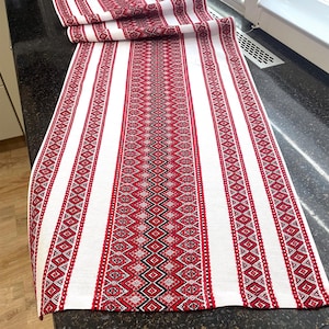 May include: A white table runner with a red geometric pattern. The runner features vertical stripes and a central design of diamonds and zigzags. The fabric is folded at the top, showcasing the pattern's repetition. The runner is approximately 152 cm long.