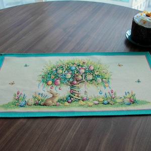 以下が含まれることがあります： ターコイズの縁取りが施された、イースターをテーマにした長方形のプレースマット。プレースマットには、装飾された木、ウサギ、卵、ヒヨコの詳細なイラストが描かれています。白いフロスティングを施した小さなケーキが皿の上にあります。