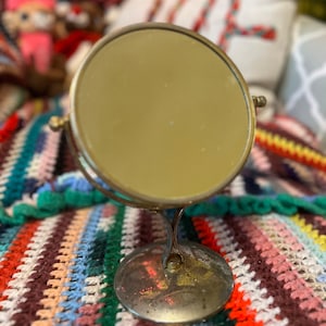 May include: A gold-toned, round, handheld mirror with a stand. The mirror is on a colourful crocheted blanket.