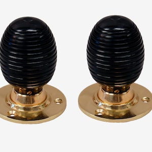 May include: Two black, ribbed door knobs with brass-colored bases. The knobs have an egg-like shape and are mounted on round plates with screw holes. The hardware is set against a plain white background.