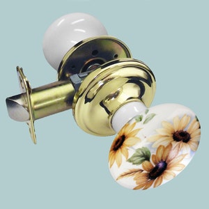 Puede incluir: Un pomo de puerta de latón con un pomo de cerámica blanca con un diseño de girasol. El pomo está unido a una manija de puerta de latón.
