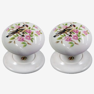 May include: Two white ceramic door knobs with a bird and floral design. Each knob features a detailed illustration of a bird perched on a branch with pink flowers and green leaves. The knobs have a round shape and a white base.