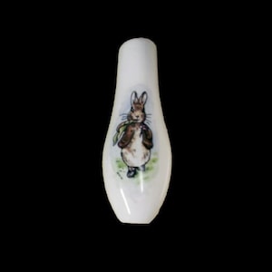 Könnte beinhalten: Ein weißer Keramik-Flaschenverschluss mit einem gedruckten Bild von Peter Rabbit, der eine Blume hält.