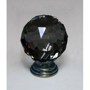 Può includere: Una sfera di cristallo grigio scuro sfaccettata poggia su una base decorativa. La sfera ha molte superfici riflettenti che catturano la luce. La base è di colore blu-grigio metallizzato con un design circolare. Questo oggetto decorativo potrebbe essere usato come fermacarte o decorazione per la casa.