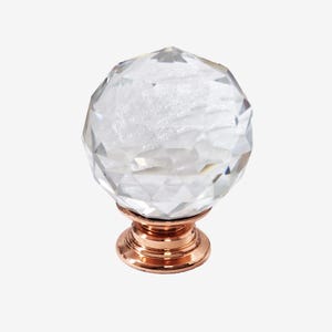 Puede incluir: Un pomo de cristal transparente y facetado con forma esférica. El pomo se asienta sobre una base de color cobre con un diseño decorativo en capas. Este herraje decorativo es adecuado para armarios y cajones.