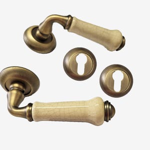May include: Antique brass door handles with cream-colored ceramic handles and matching keyhole covers. The hardware has a vintage aesthetic, with a warm, aged finish. The ceramic handles have a crackle glaze.