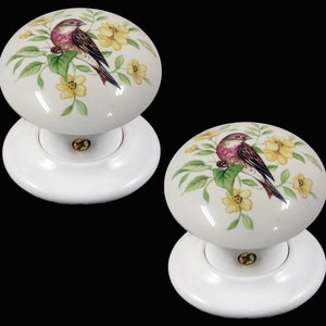 Peut inclure: Deux boutons de porte en céramique blanche avec un motif floral représentant un petit oiseau aux plumes brunes et roses perché sur une feuille verte. Les fleurs sont jaunes avec des centres blancs.