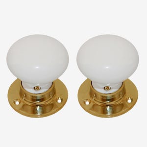 May include: Two white ceramic door knobs with a round shape, mounted on a gold-coloured metal base. The base has a circular shape with screw holes. The knobs are a classic design.