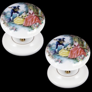 Peut inclure: Deux boutons de porte en céramique blanche avec un motif floral et une scène de personnes dans un jardin. La scène comprend un homme jouant de la guitare et deux femmes.