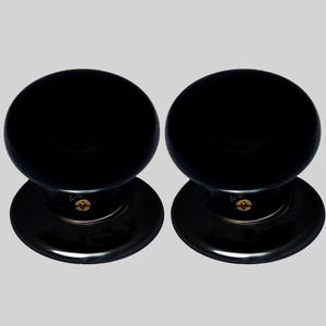 May include: Two black ceramic door knobs with a round shape and a flat base. Each knob has a small gold screw in the centre.