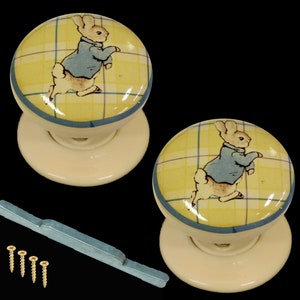 May include: Two ceramic door knobs with a yellow and blue checked background and a white rabbit wearing a blue jacket. The knobs are attached to white bases.