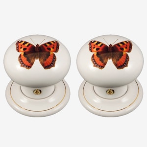 May include: Two white ceramic door knobs with a gold trim. Each knob features a colorful butterfly design.