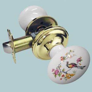 Może przedstawiać: Biała porcelanowa gałka drzwiowa z kwiatowym wzorem i złotą metalową podstawą. Gałka ma wzór ptaka i kwiatów.