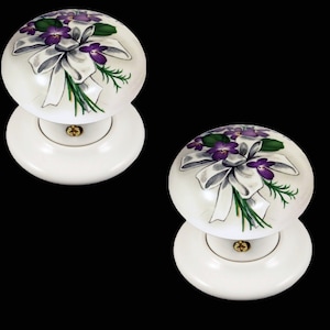 May include: Two white ceramic door knobs with a floral design. The knobs feature a bouquet of purple flowers with white ribbon and green leaves.